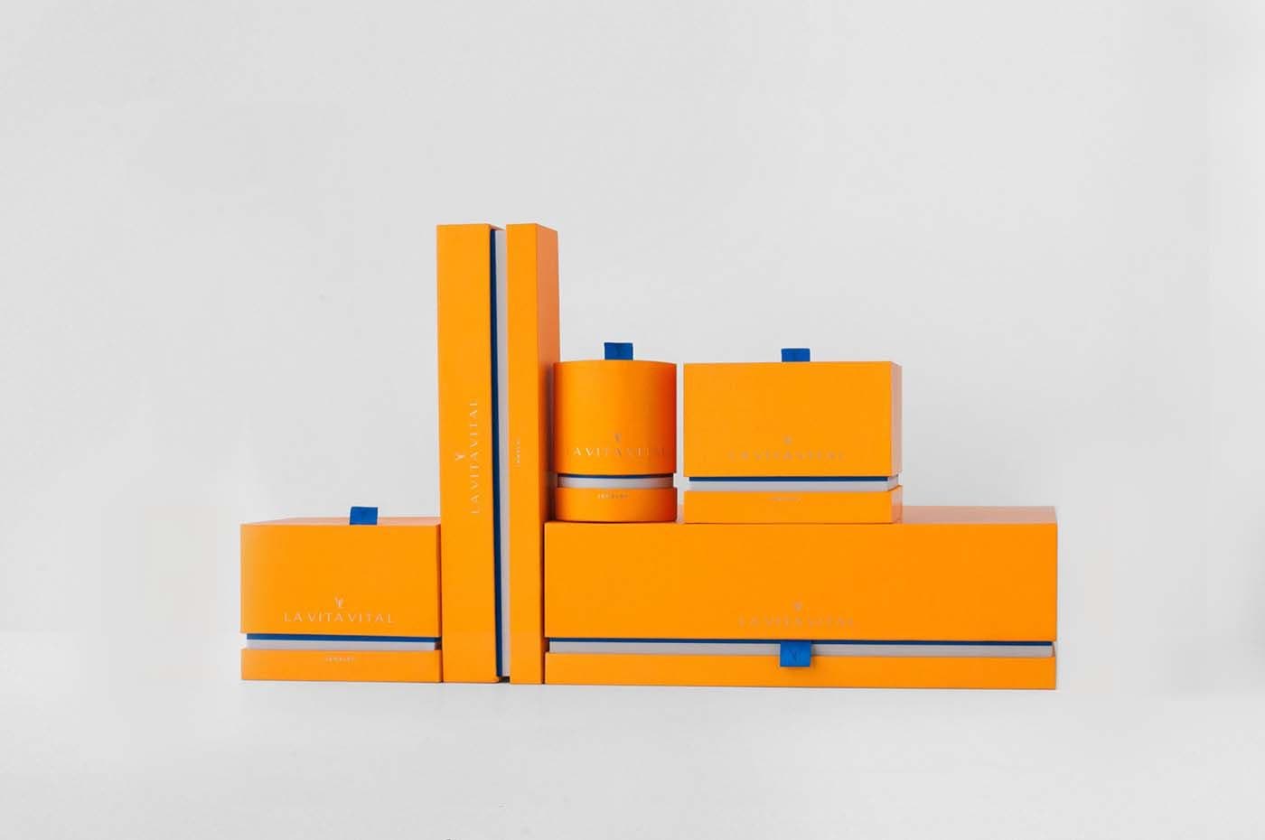 Overhead view of orange La Vita Vital jewelry packaging boxes and a book-style case arranged in an angular cluster, with blue tab pulls on a white background.