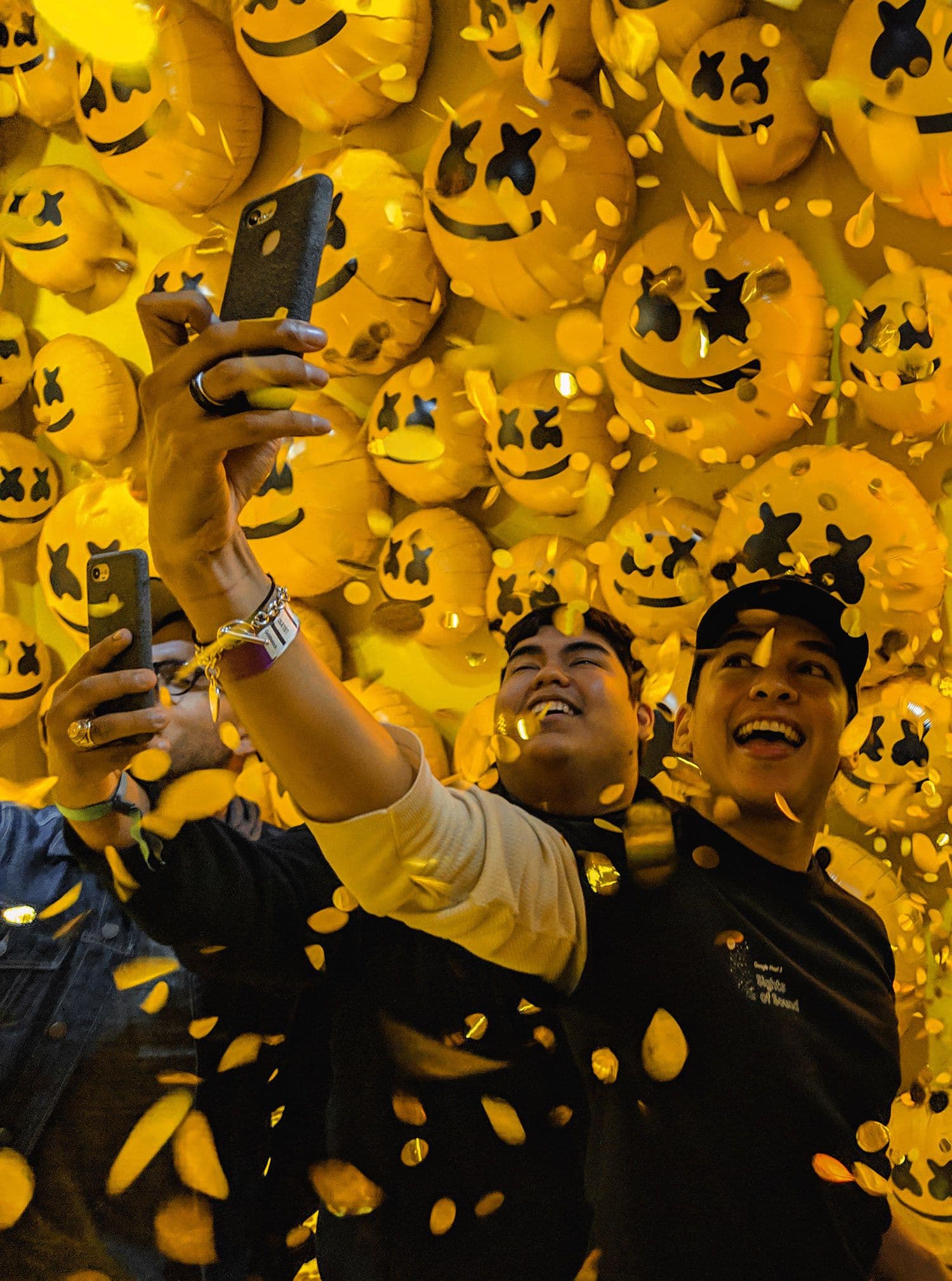 Two people taking a selfie surrounded by a wall of glowing orange emoji face sculptures.