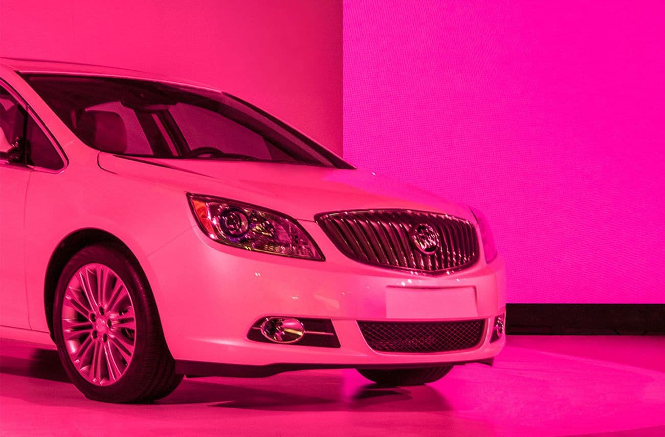 Front three-quarter view of a white Buick sedan lit in pink light.