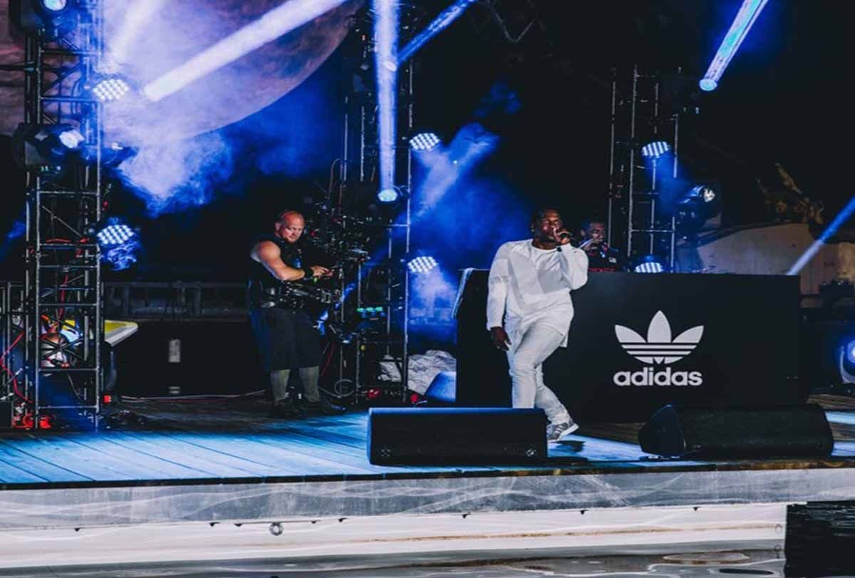 Performer in white singing on stage beside a cameraman, with an adidas trefoil logo on a speaker stack and blue stage lighting.