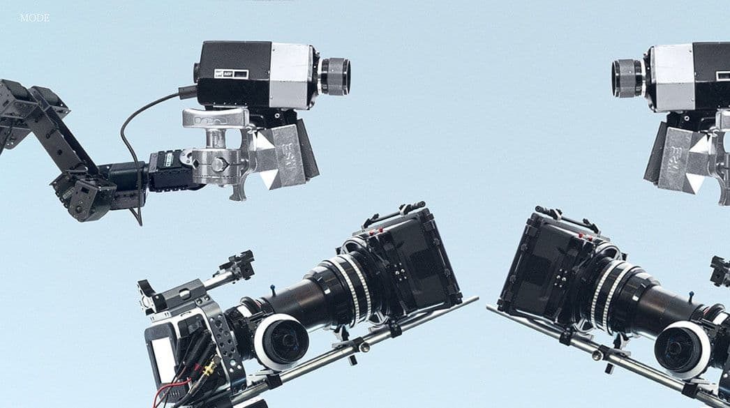 Close-up of robotic camera rig arms and film cameras crossing against a pale blue backdrop, no model in frame