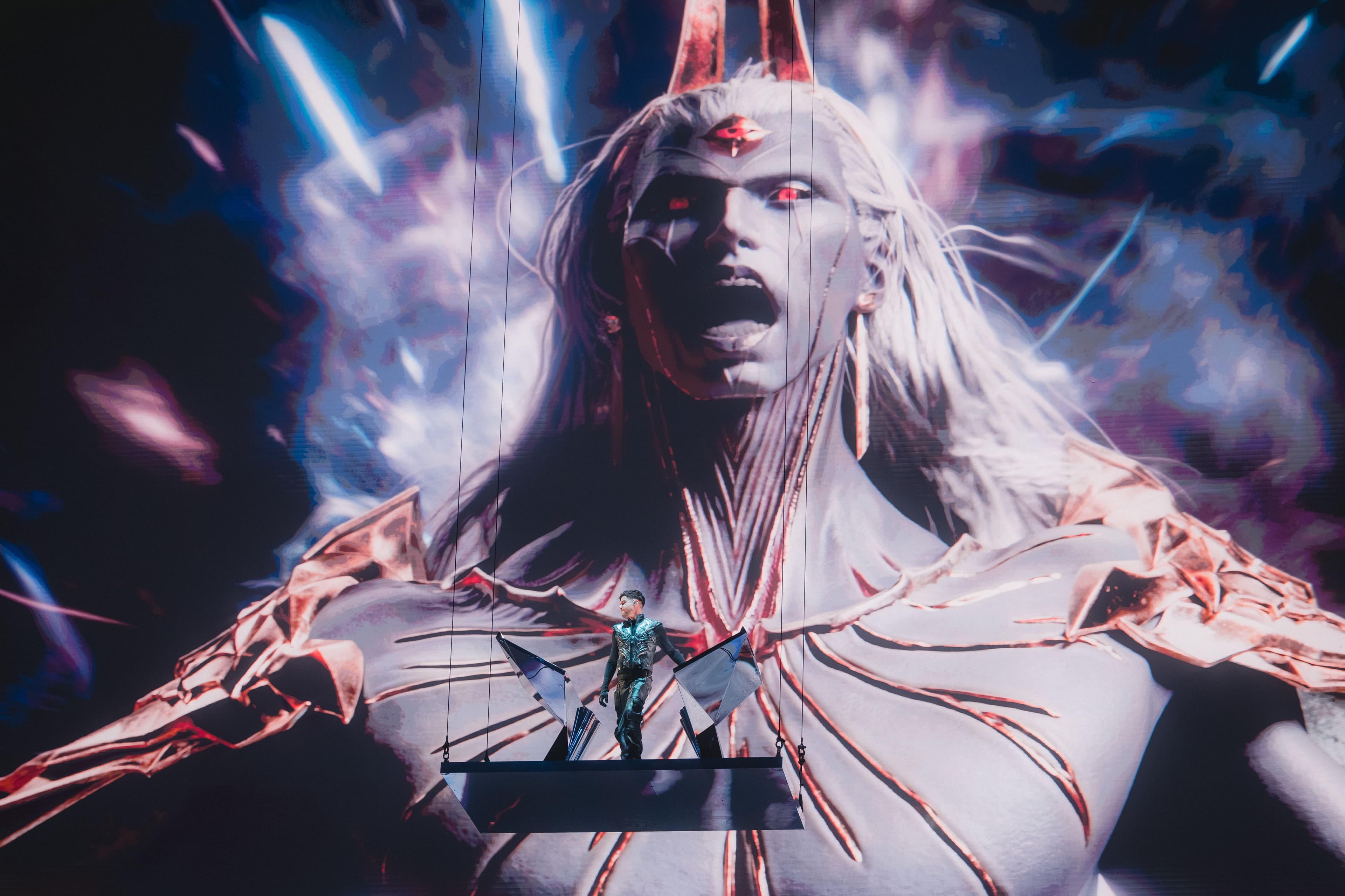 Performer with long white hair and a flowing cape, arms outstretched, lit in red light.