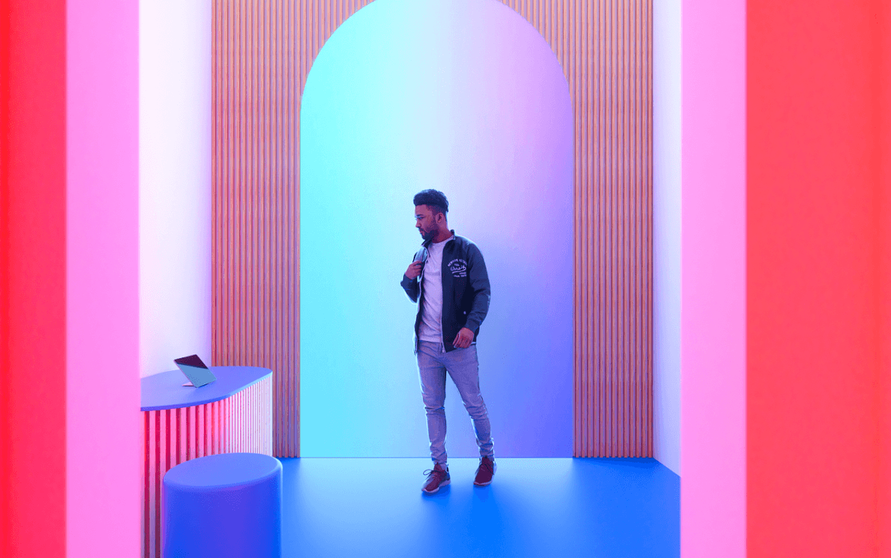 A man in a bomber jacket stands under an arched doorway lit in blue and pink, next to a low pedestal with a phone on it.