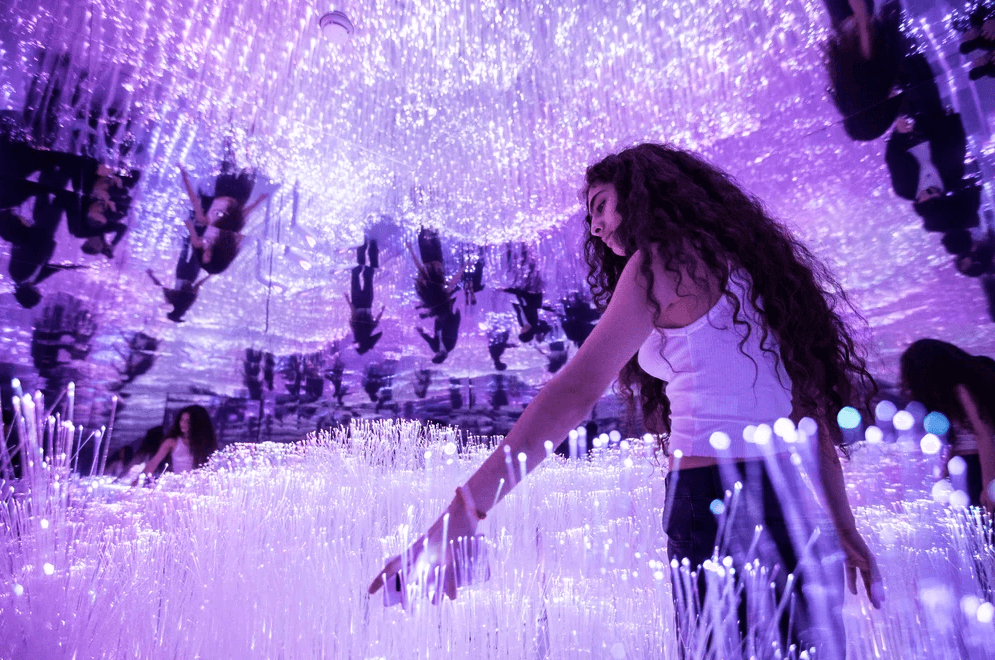 A person reaches out to touch hanging purple fiber-optic strands while walking through a lit installation.