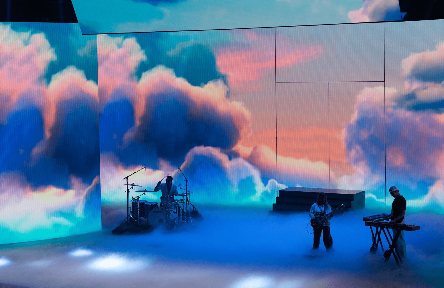 Musicians performing on a foggy stage beneath screens showing orange cloud imagery