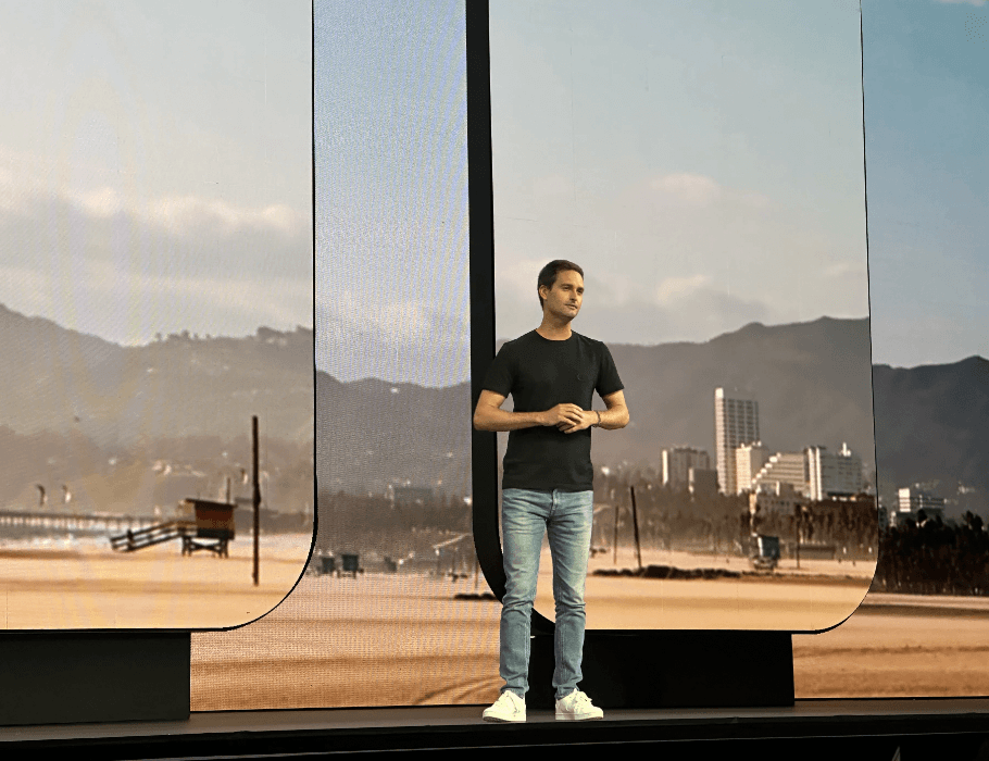 Presenter on stage in front of a large window overlooking mountains at dusk.