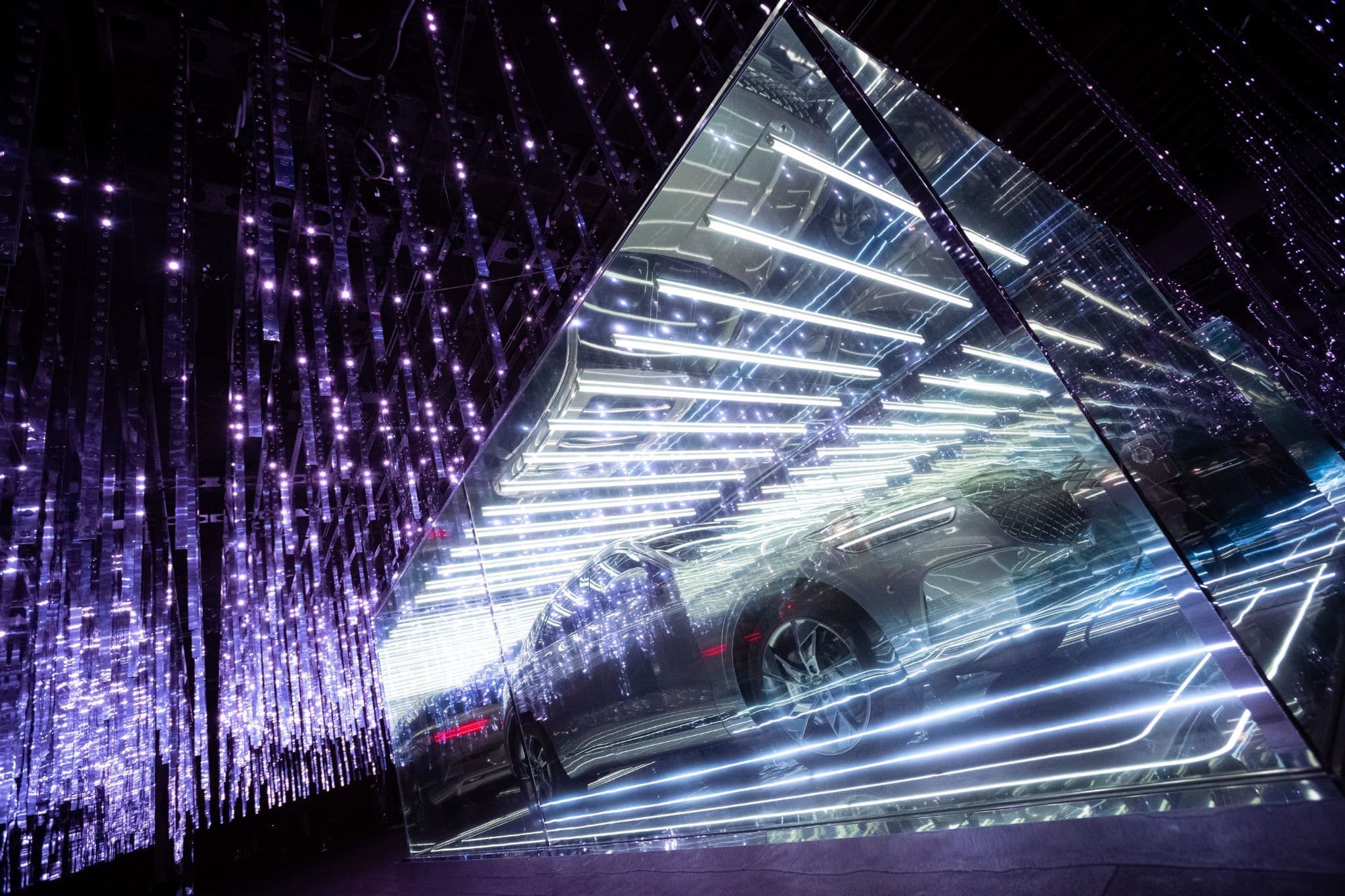 A car reflected inside a triangular mirrored prism structure lit with violet string lights and white neon lines.