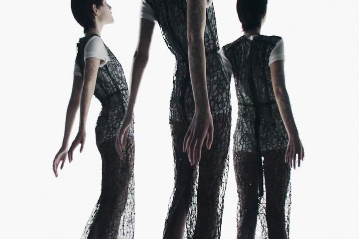 Three models in profile against a white backdrop, wearing black openwork knit gowns over white tops, arms hanging loosely.