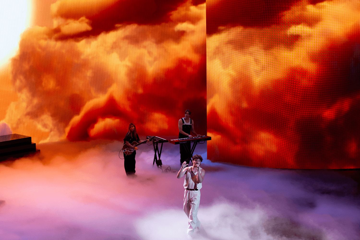 Performers on a foggy stage beneath large screens showing orange cloud imagery