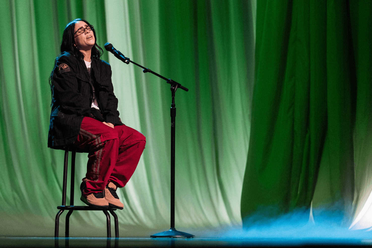 Performer seated on a stool singing against a green-lit curtain backdrop