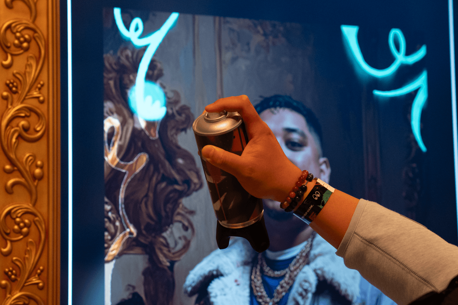 A close-up of a hand spraying blue paint onto a gold-framed screen displaying a glowing portrait.