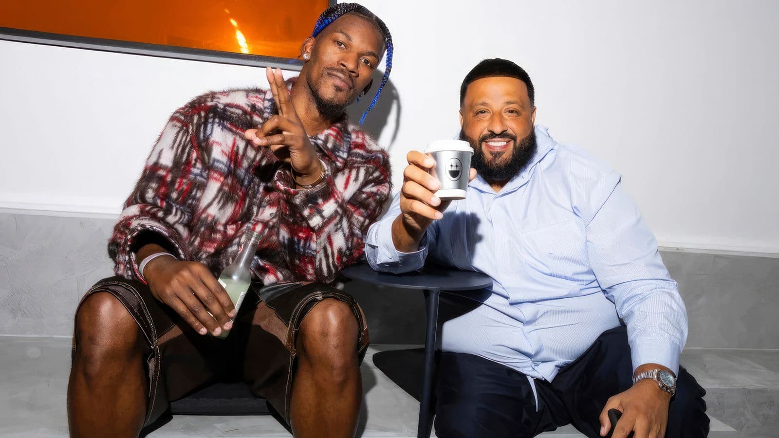Two men sitting outdoors, one making a hand gesture while holding a drink, the other holding a cup