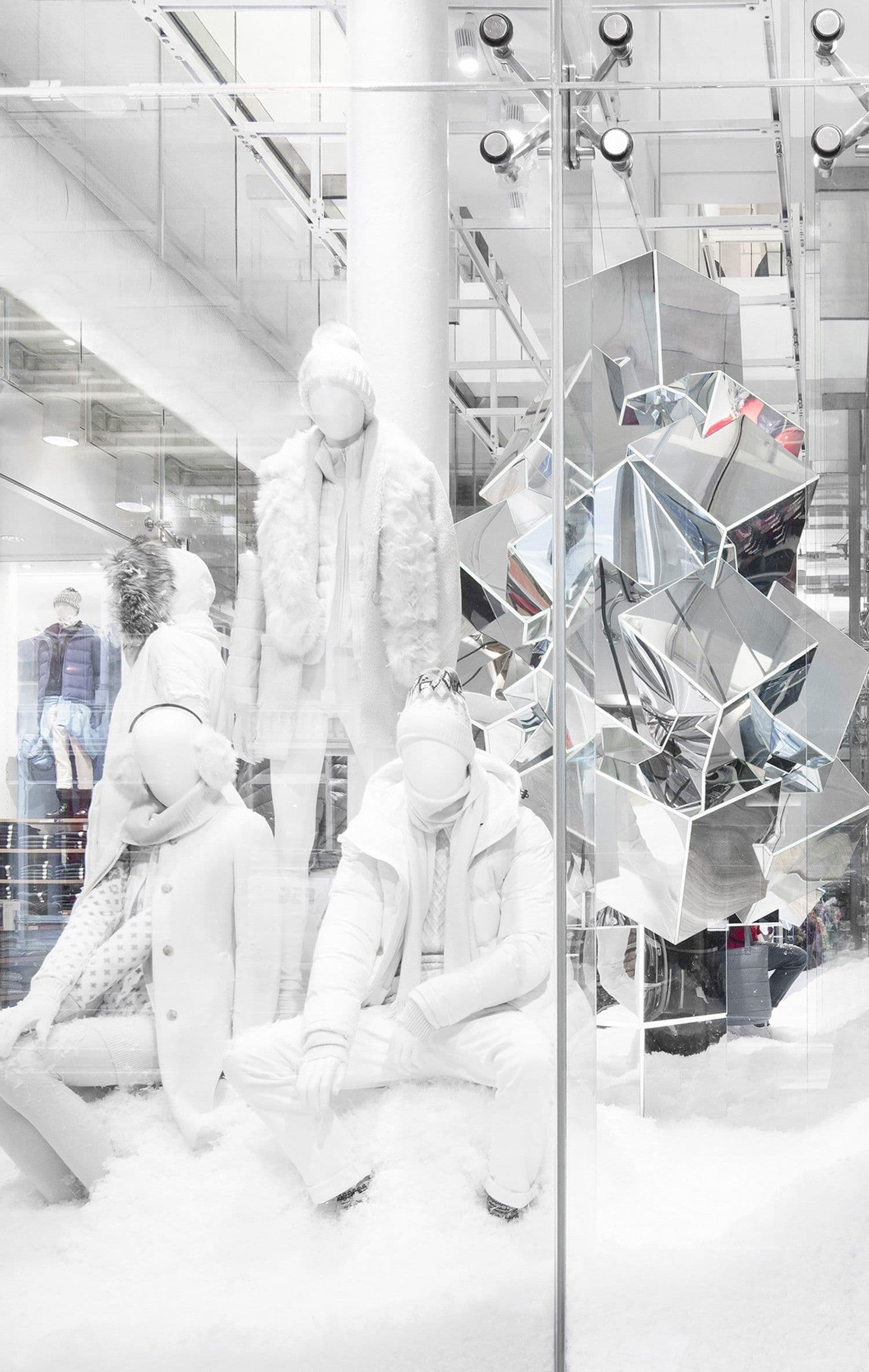 All-white mannequin display of people in winter coats and beanies seated in fake snow, beside a faceted mirrored sculpture behind glass.