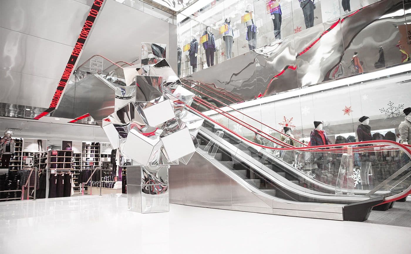 Faceted mirrored sculpture beside an escalator, reflecting a wall of screens showing models in winter outerwear.