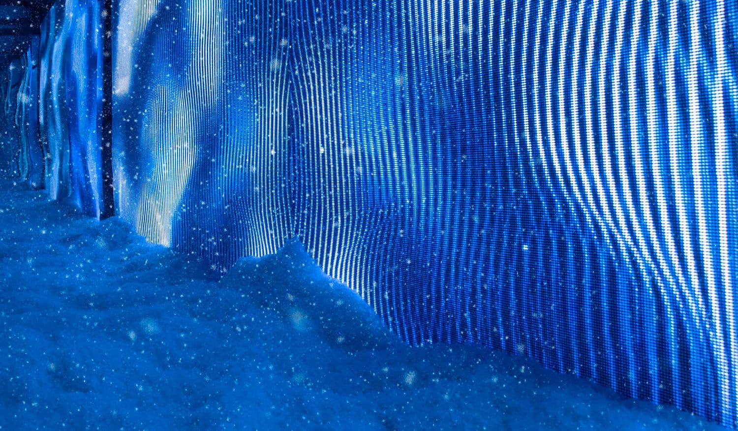 Blue light installation with a rippling wave pattern beside a textured, snow-like wall.
