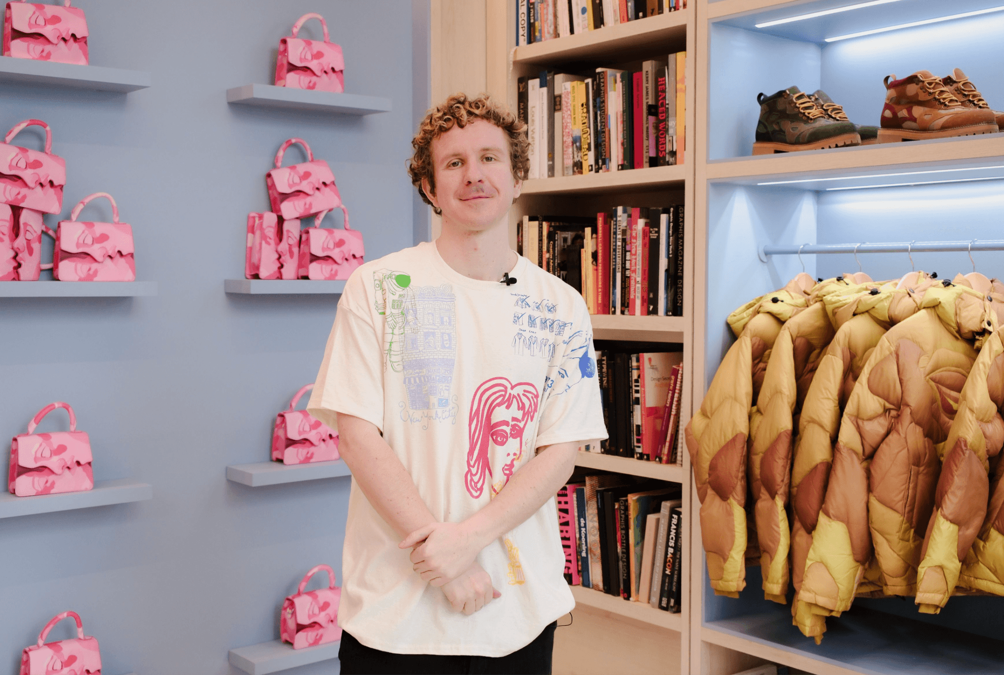 Man in a printed graphic t-shirt standing in a store aisle with pink handbags on wall shelving and shoes displayed behind him