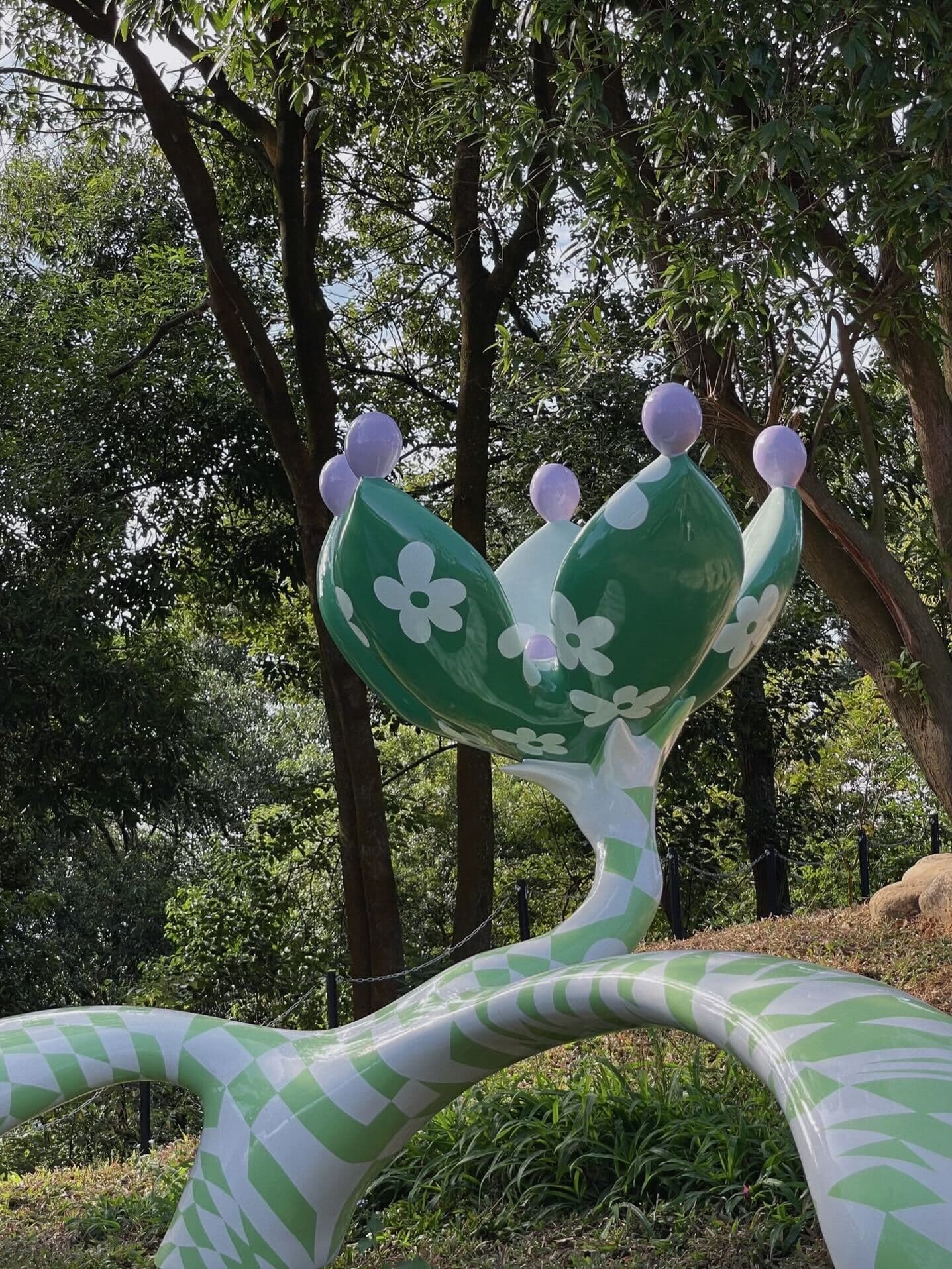 Green ceramic sculpture shaped like flowering cactus paddles with lavender ball tips, set among trees.