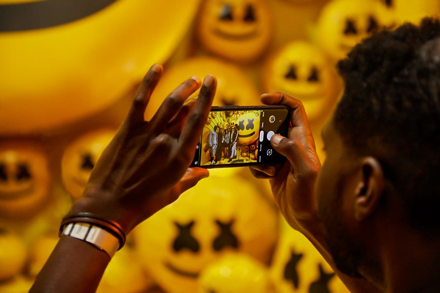 Hands holding a phone to photograph a glowing orange emoji sculpture surrounded by smaller emoji orbs.