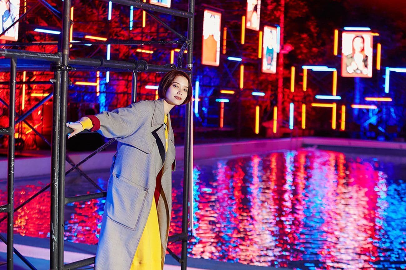 Woman in a long coat leaning against an illuminated metal frame beside a reflecting pool lit with colorful lights.