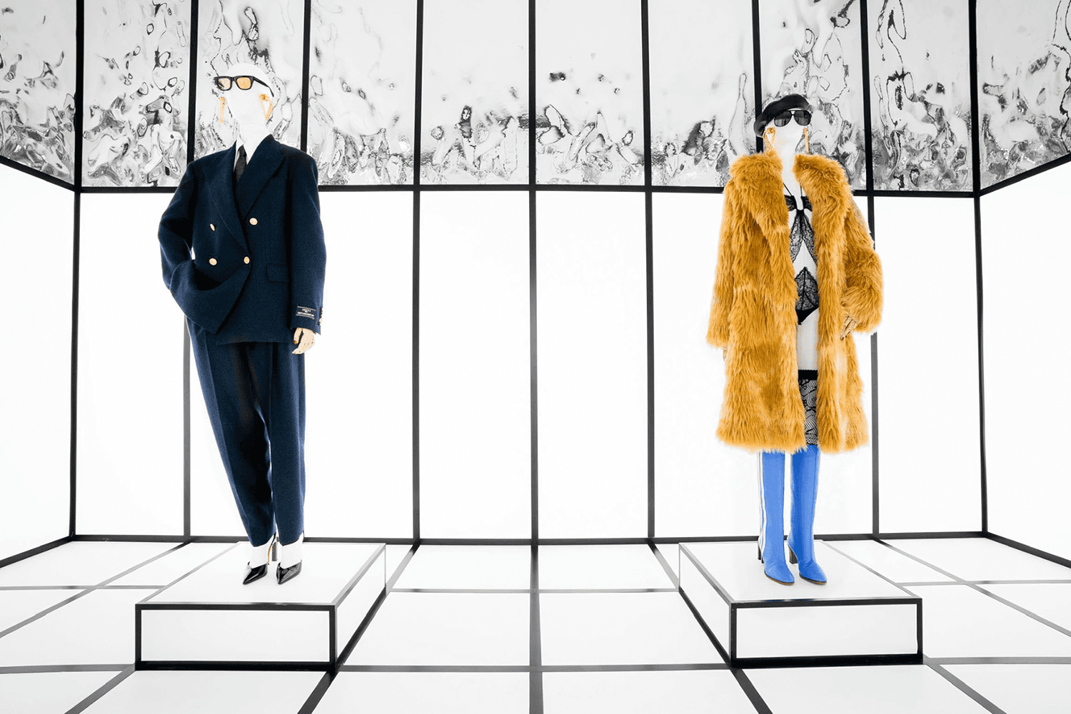 Two mannequins, in a navy uniform-style look and a mustard fur coat, inside a black-and-white striped glass enclosure.
