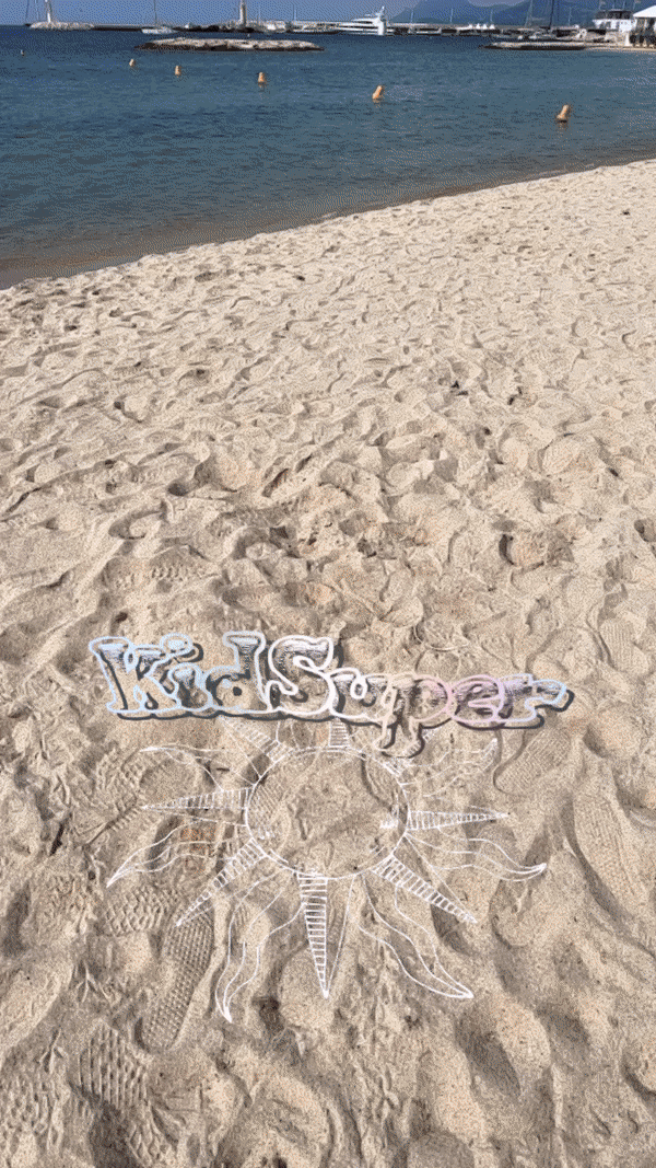 A sandy beach shoreline with the word 'KidSuper' and a sun design drawn in the sand, boats visible on the water beyond.