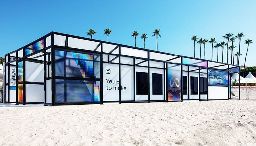 A glass-walled pavilion on the beach with an Instagram logo and the text 'Yours to Make', palm trees lining the background.