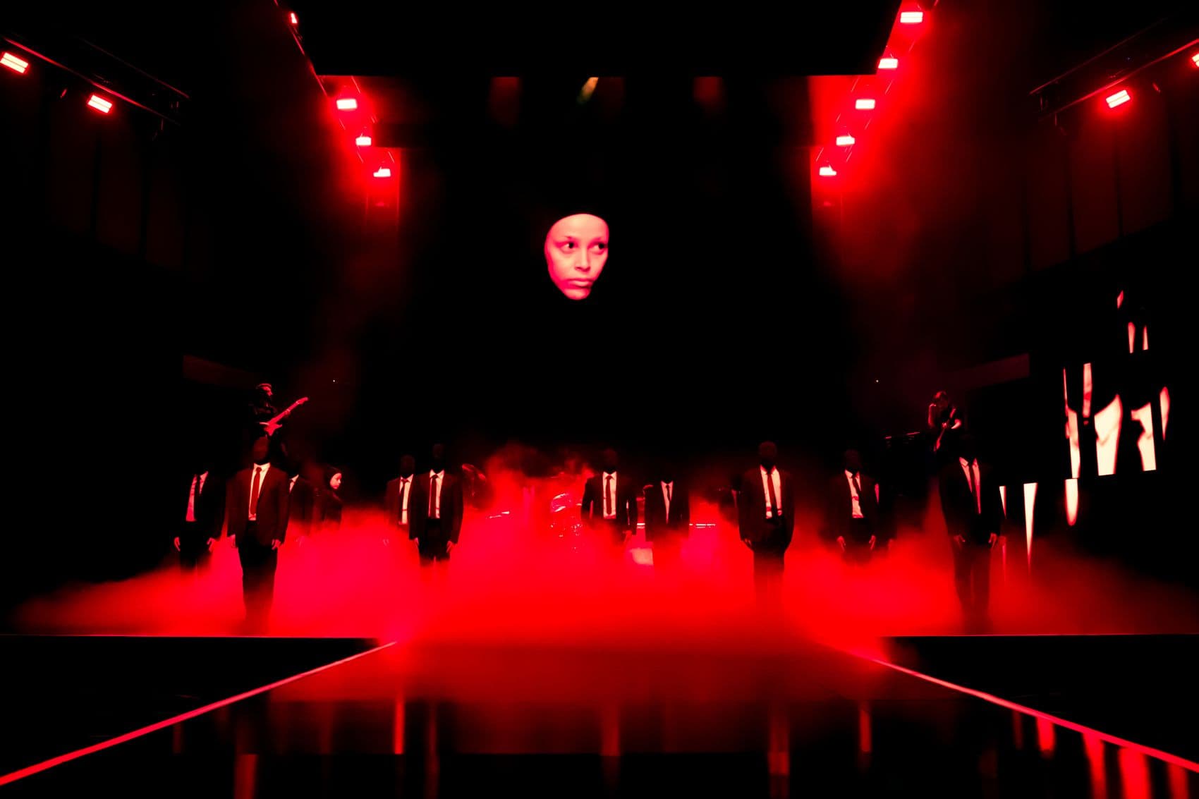 Close red-lit shot of a performer standing at a lectern-like set piece flanked by angular panels.