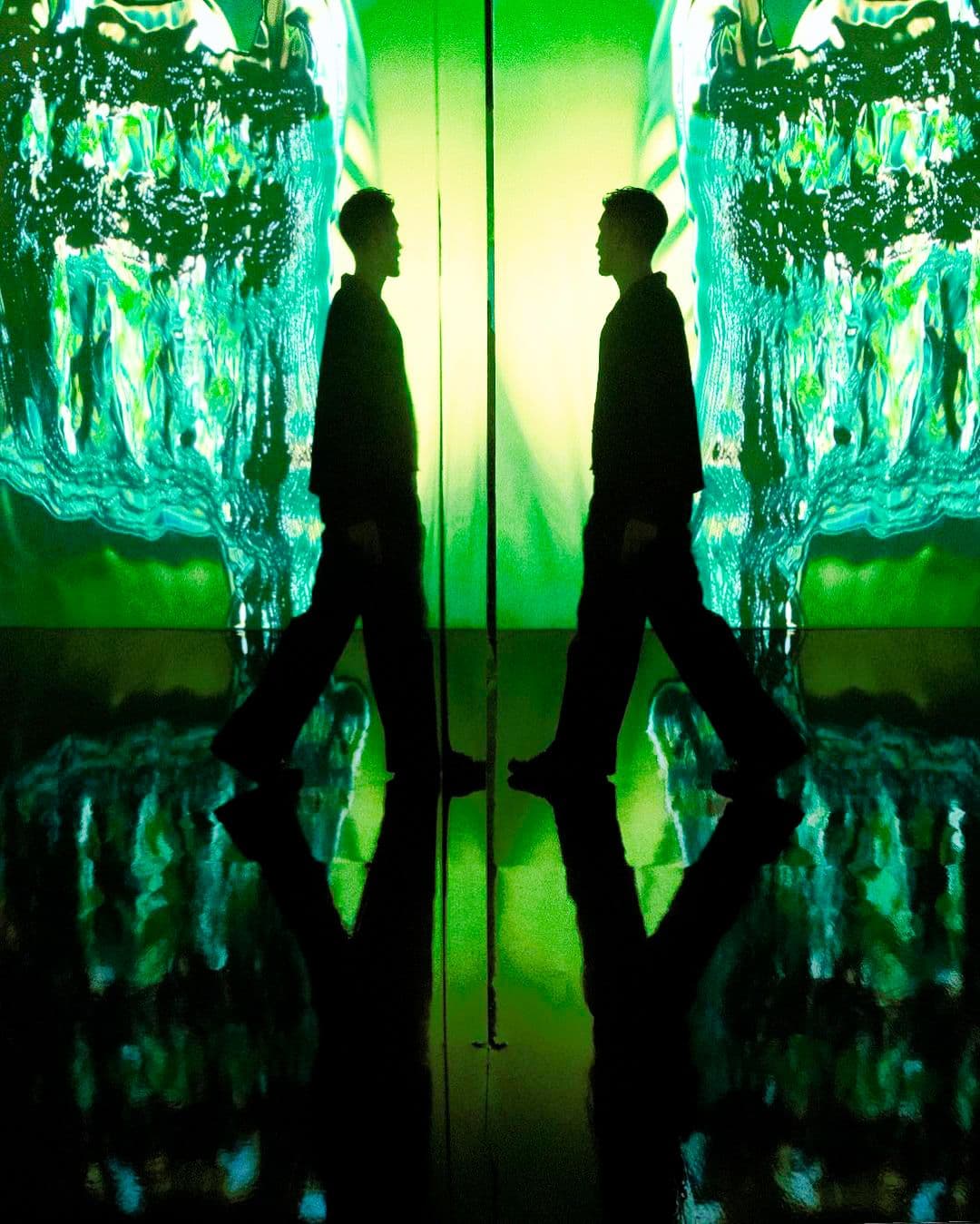 Two silhouetted figures walking through a green-lit glass corridor, reflected on a glossy floor.