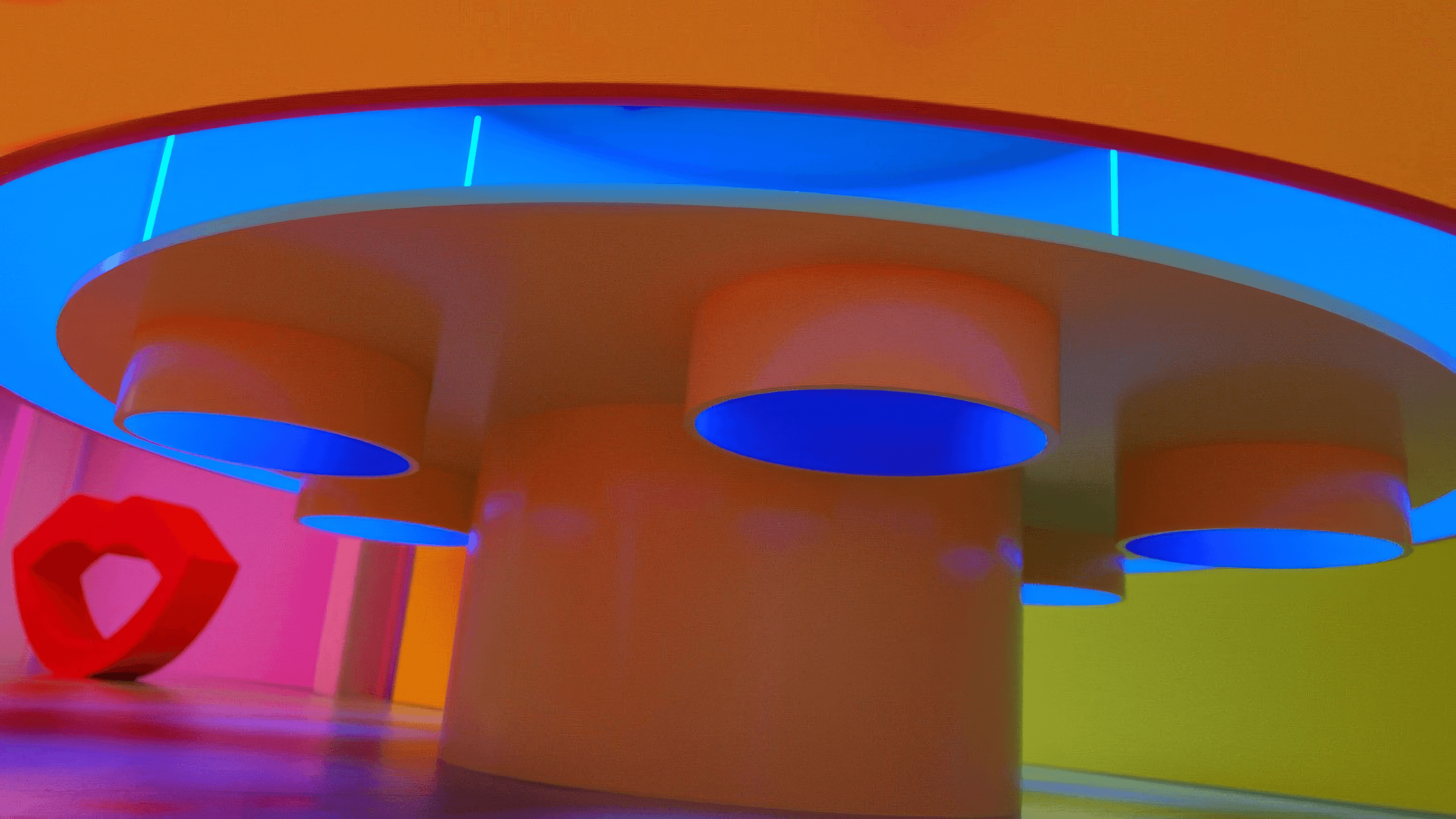 Wide shot of an orange pop-art exhibition space with round pedestal tables under a blue ceiling and a red heart-shaped sculpture.