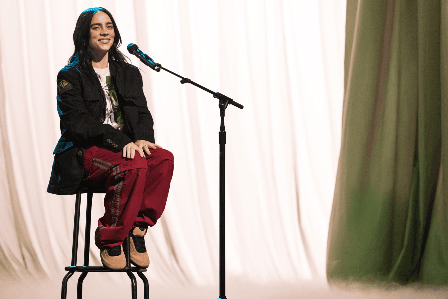 Performer seated on a stool singing into a microphone against a soft cream curtain backdrop