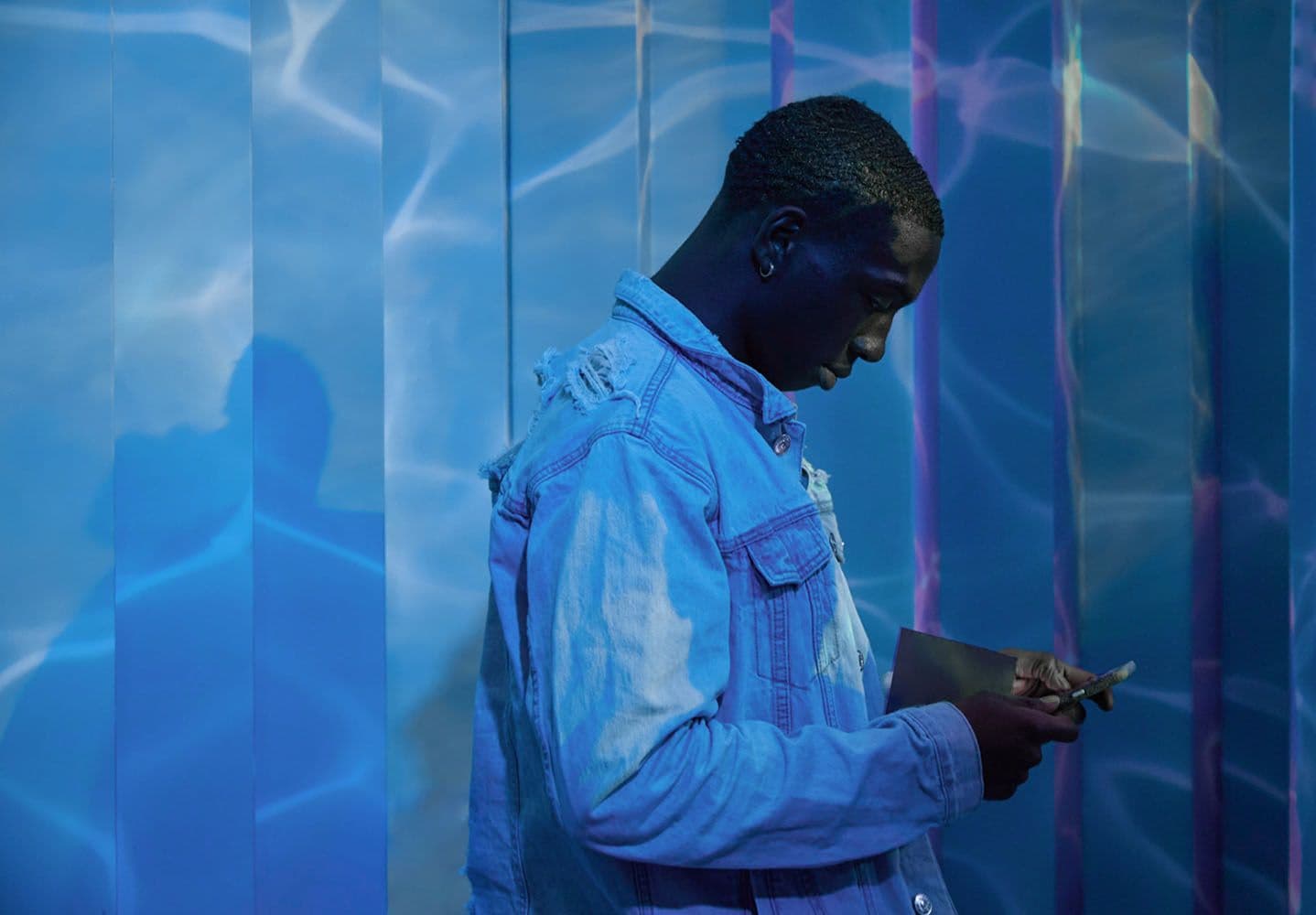 A man in a denim jacket looking at his phone against a blue-lit striped wall.