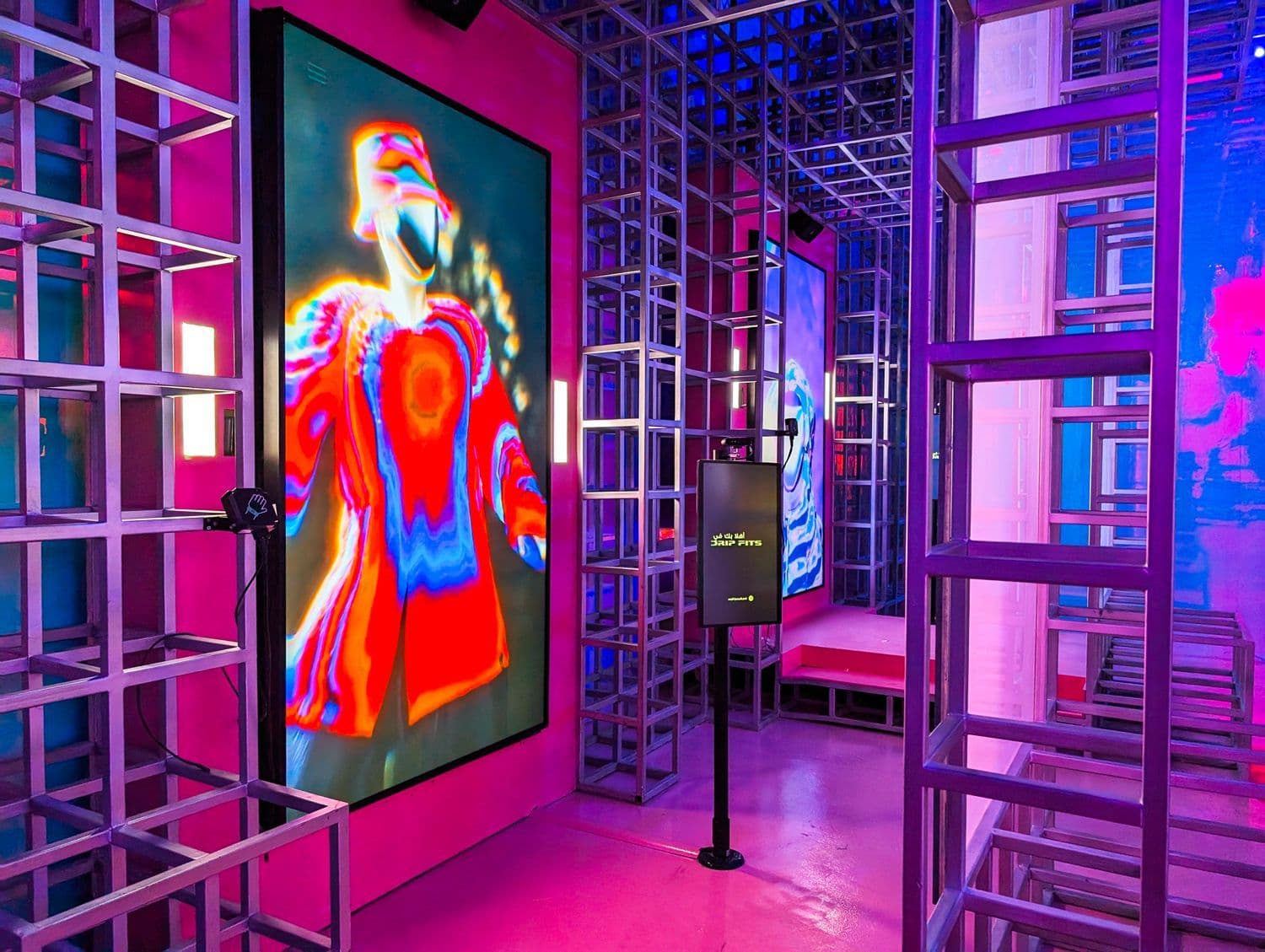 A red-lit corridor lined with metal scaffolding towers, with an LED screen showing an animated red humanoid figure.
