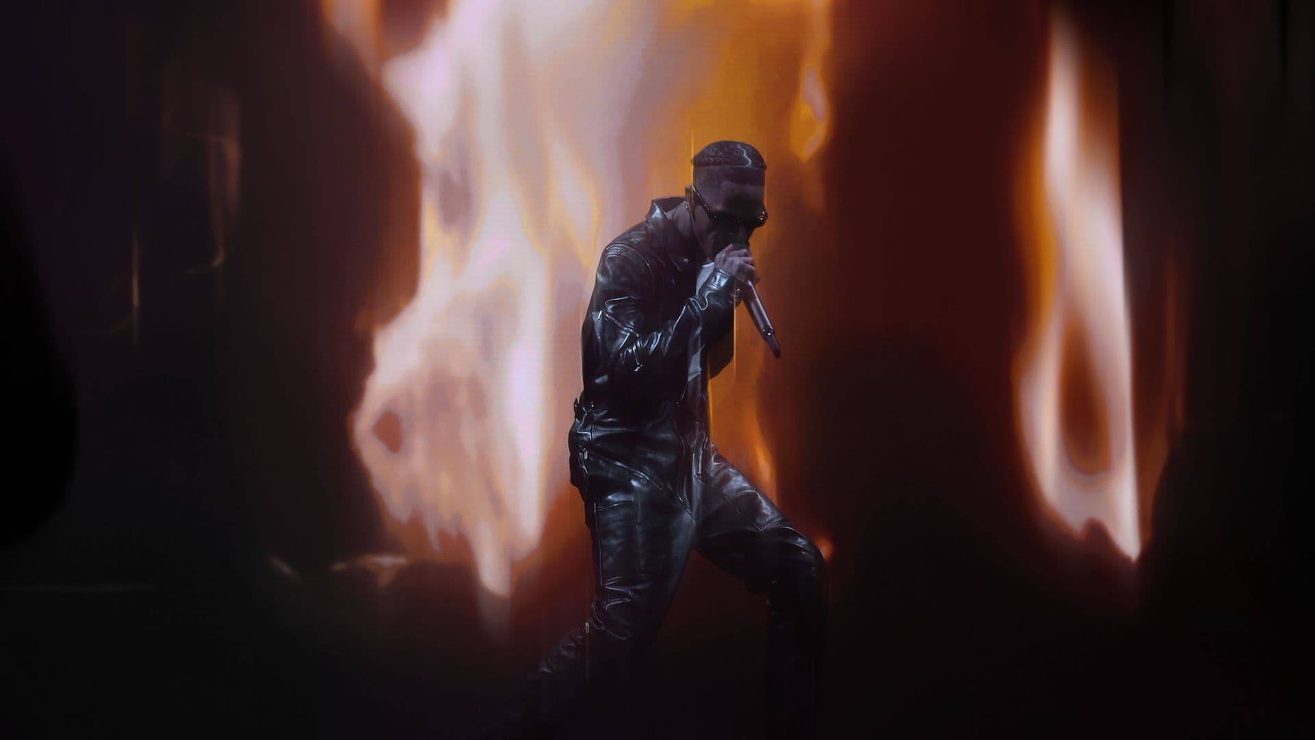 Performer bent forward singing into a microphone, lit from behind by orange stage flames.