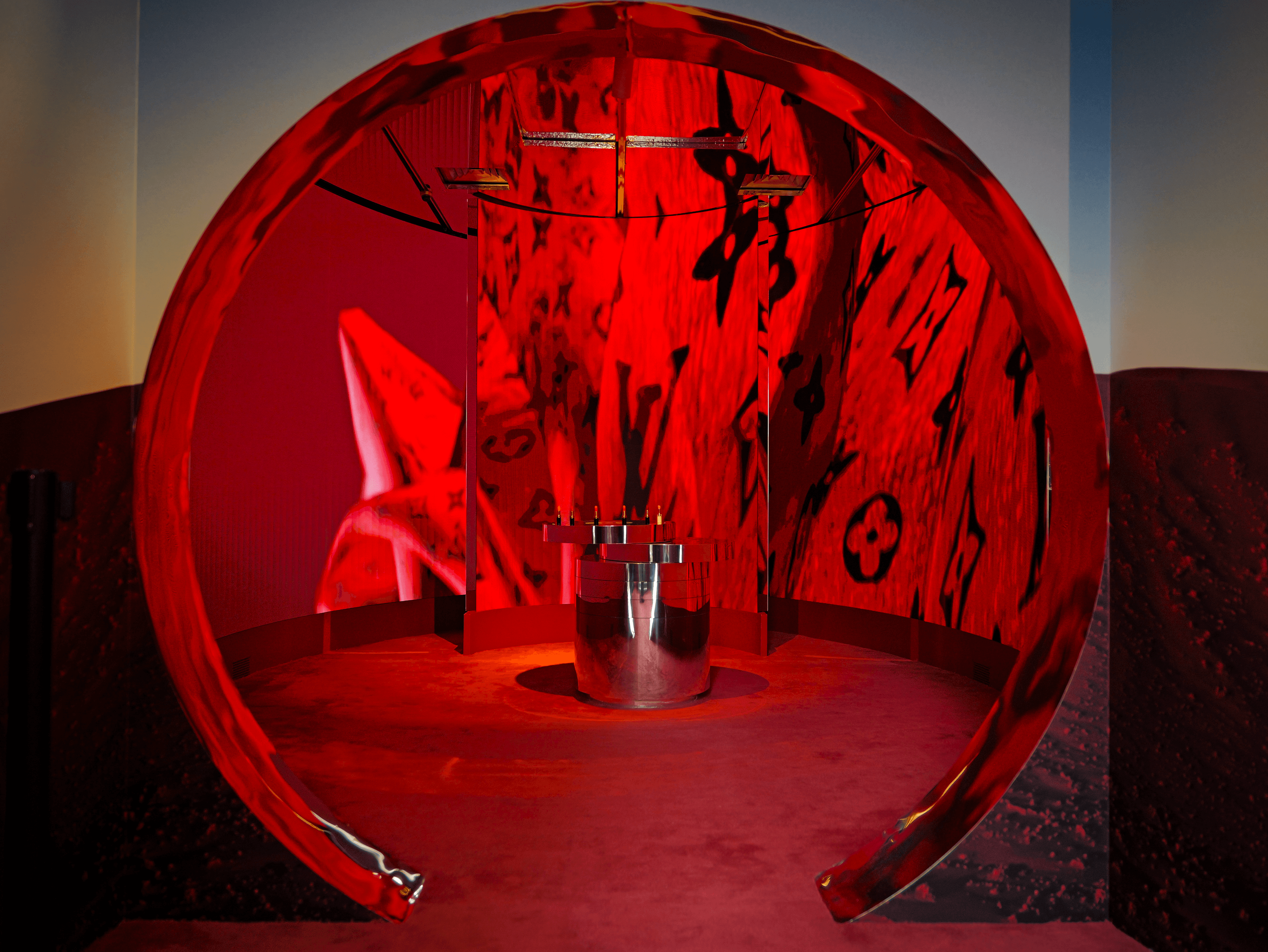 Red circular tunnel with the Louis Vuitton monogram pattern displayed in black on cross-shaped screens, cylindrical plinth at center.