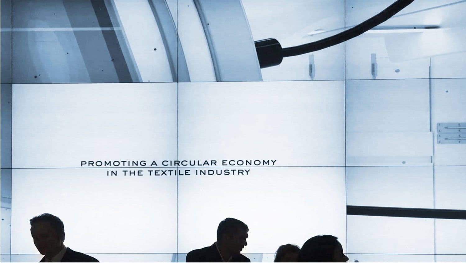 Illuminated screen reading "Promoting a circular economy in the textile industry" above silhouetted attendees.