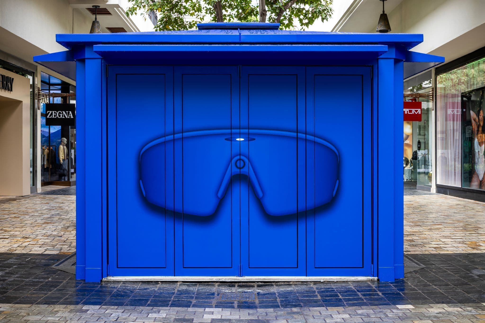 Closed blue kiosk with a large sunglasses graphic printed across its front panels, set in an outdoor shopping plaza.