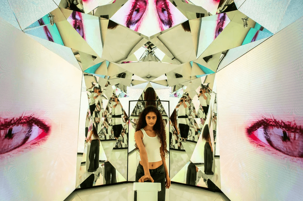 A woman stands inside a mirrored tunnel reflecting a repeated red lips and eye graphic pattern.