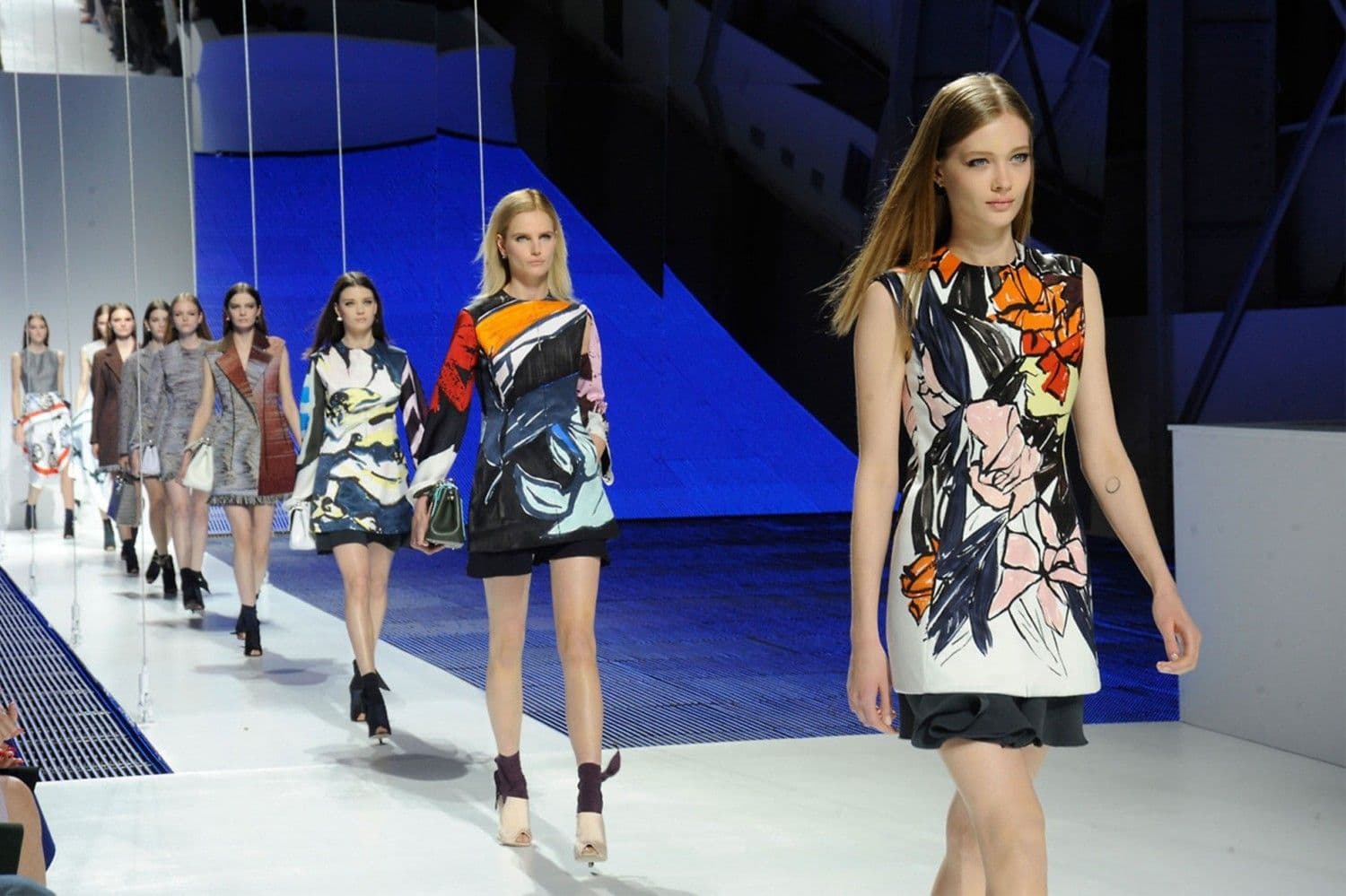 Two models walk down a tiered runway in colorful abstract-print dresses, with rainbow gradient panels in the background.