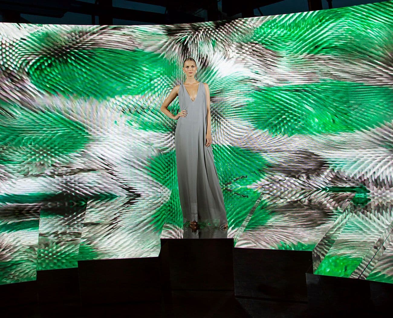 A model in a grey gown standing on a pedestal in front of a curved LED screen displaying a green fern-like pattern.