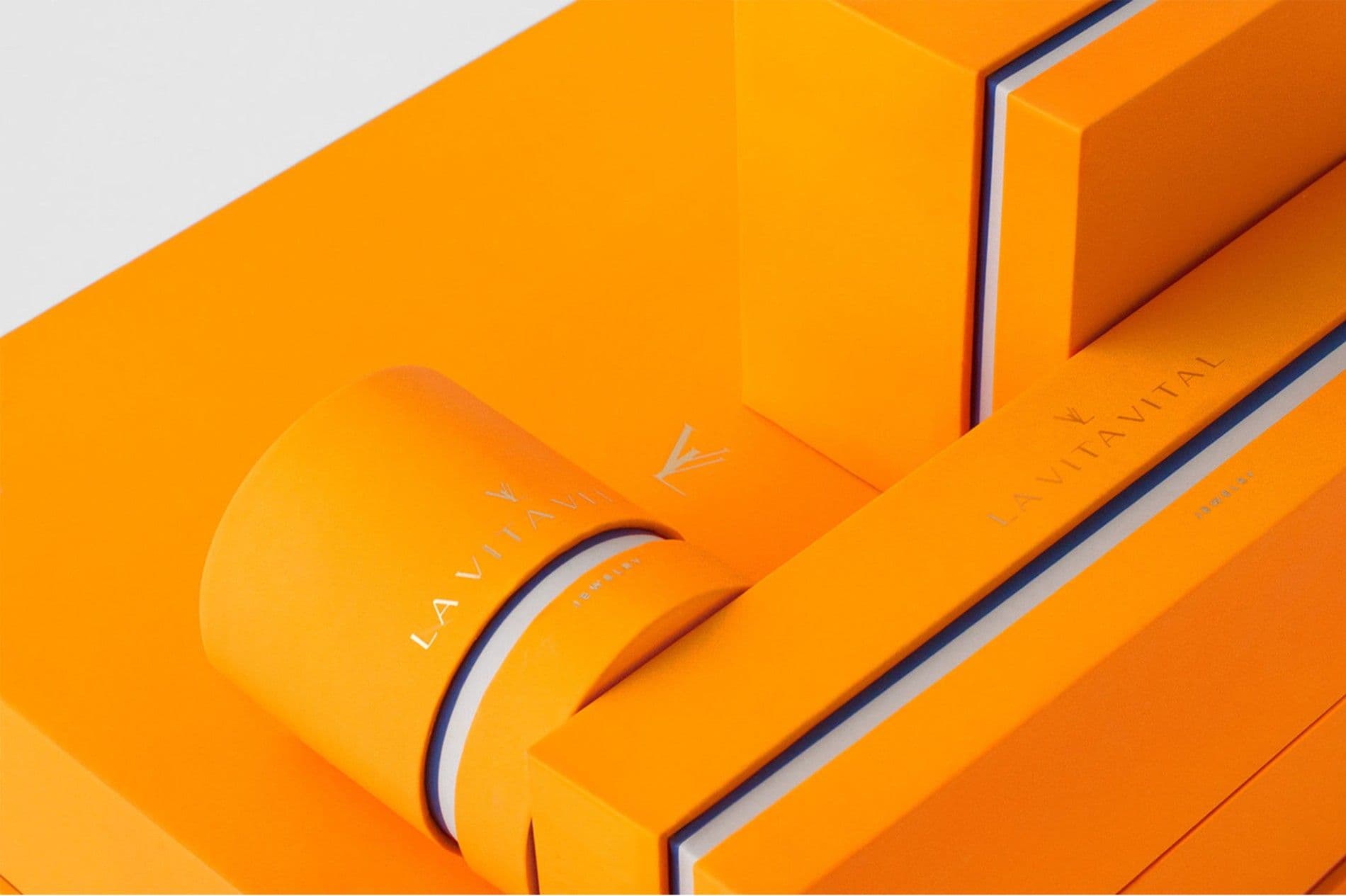 Close-up of a cylindrical orange box lid embossed with the La Vita Vital wordmark and logo, set against a taller orange case with a navy edge stripe.
