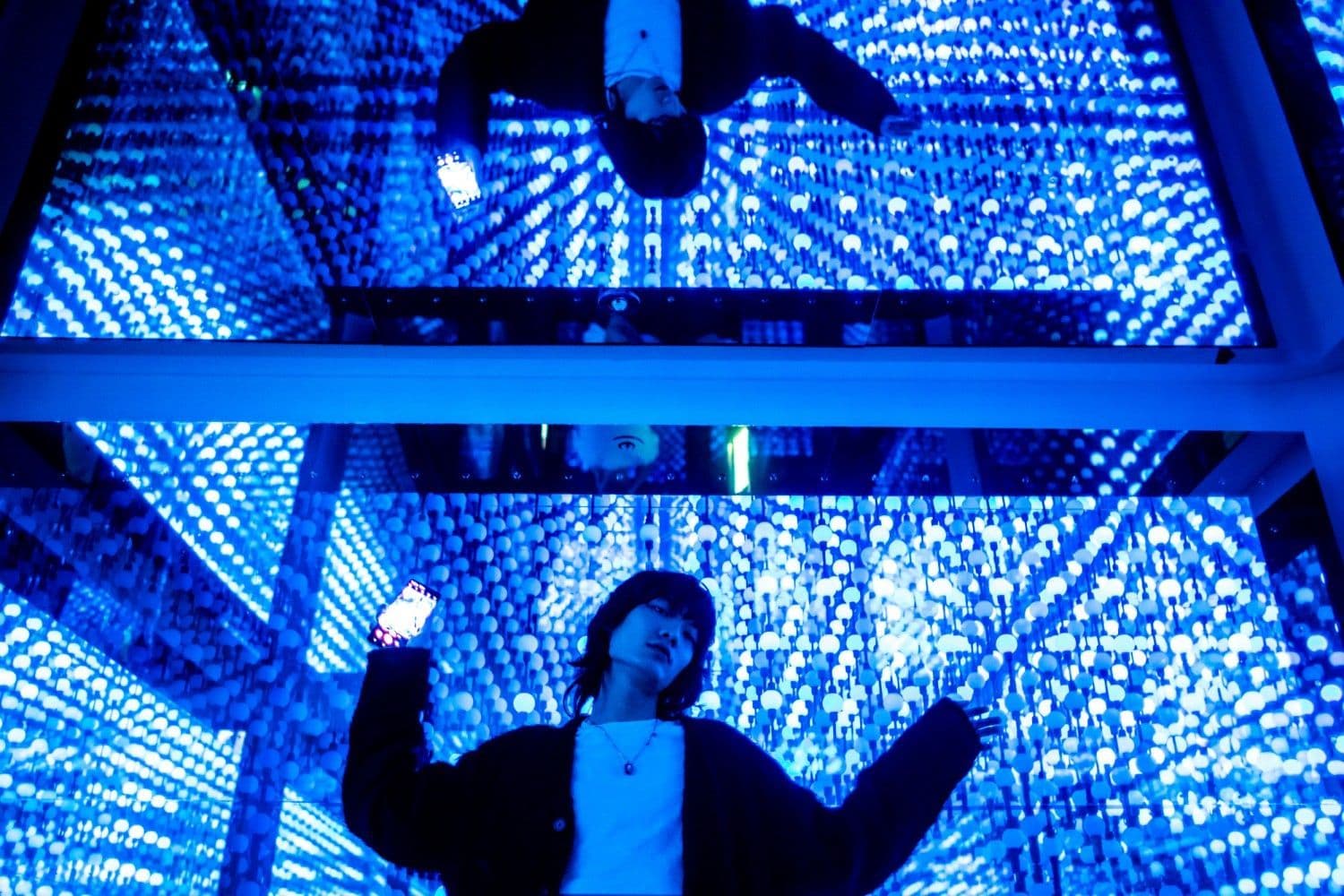Aerial view looking down into a blue LED-lit mirrored maze as a person raises their arms below.