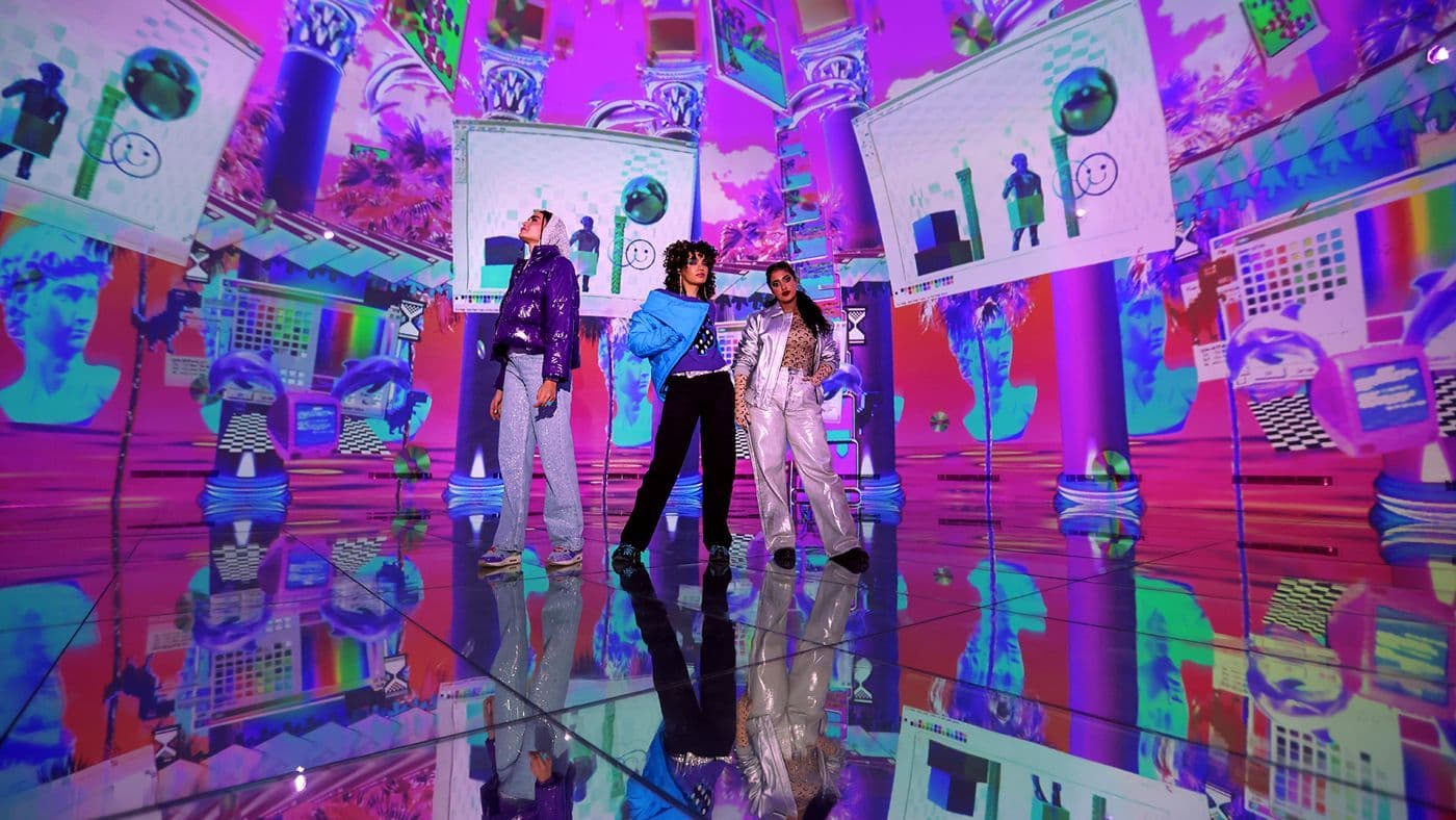 Three people stand on a mirrored floor inside an immersive room with walls of colorful, glitch-patterned video screens.
