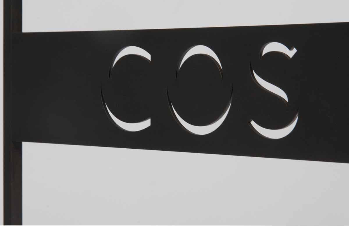 Close-up of the COS wordmark cut out of a black metal panel.
