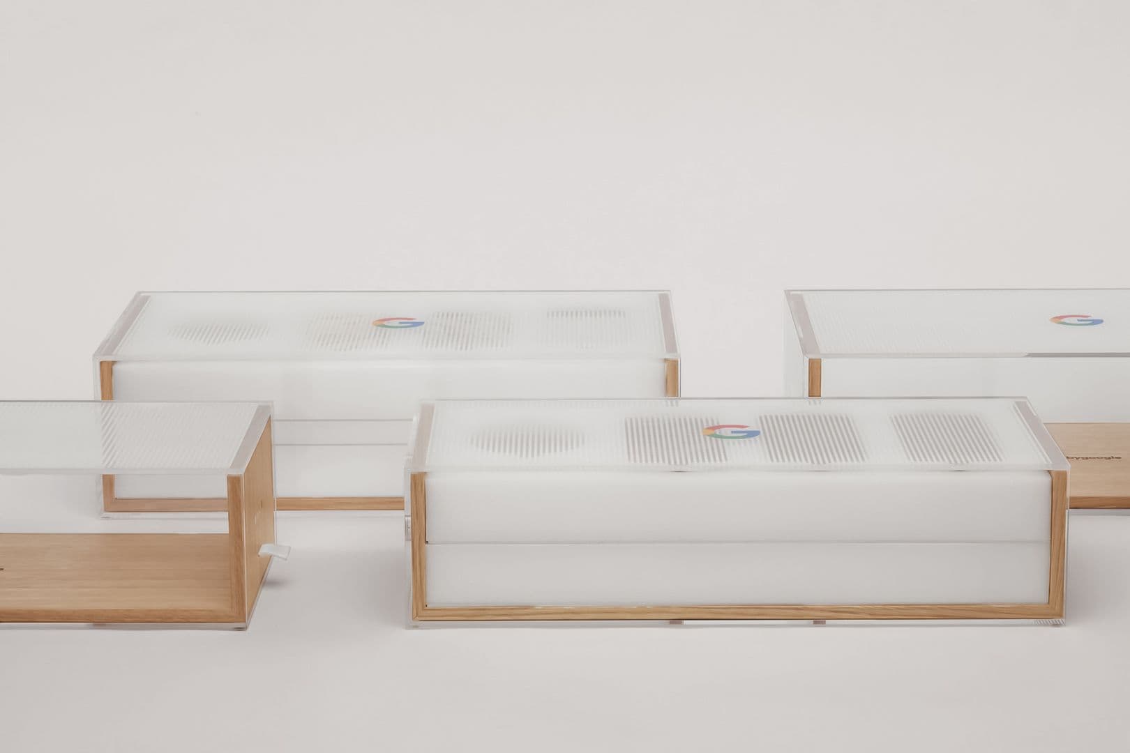 Several frosted acrylic and wood packaging boxes with perforated speaker-grille lids, each stamped with a small Google logo, arranged in a row.