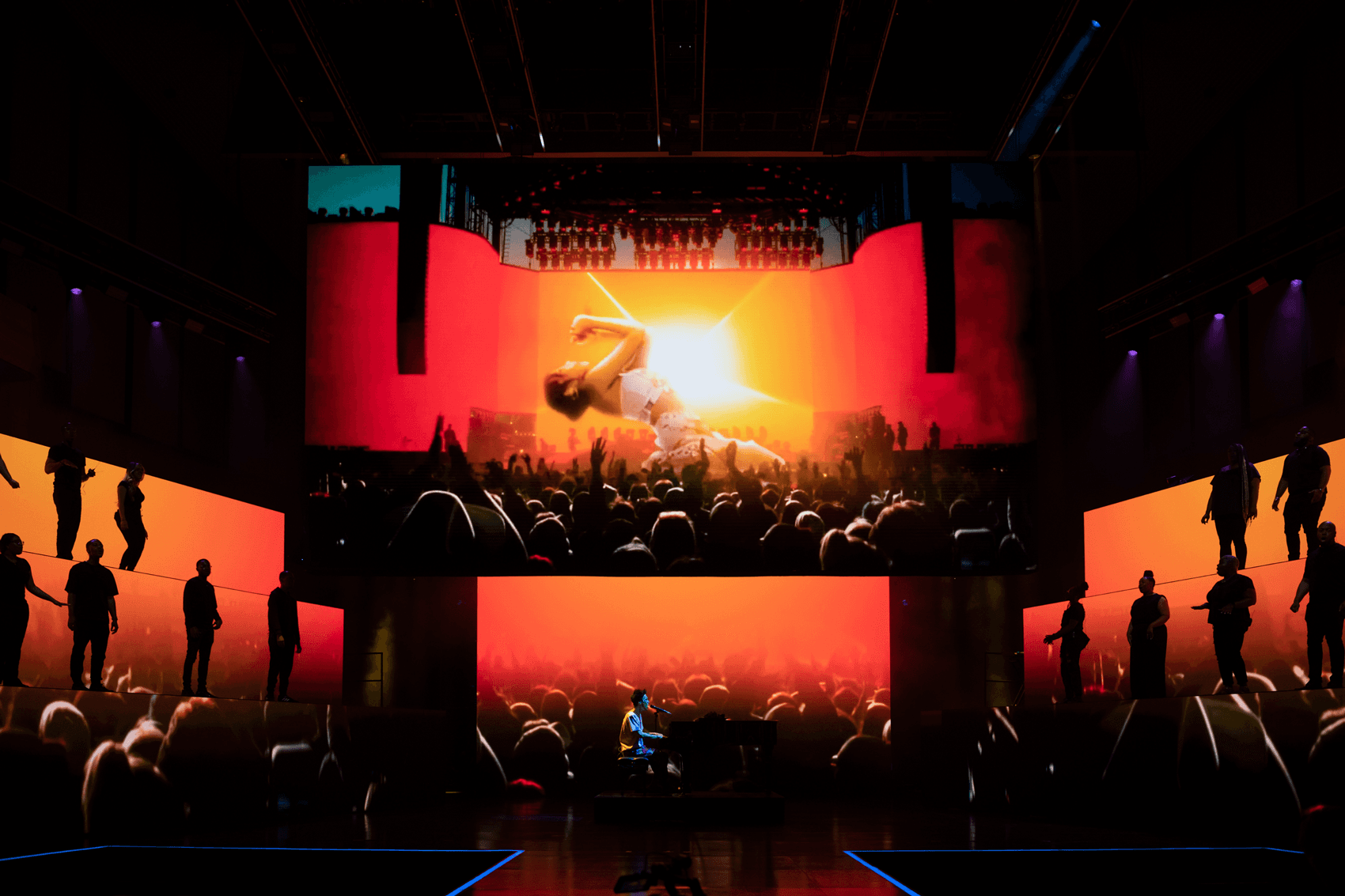 Wide stage view with an orange sunset screen above silhouetted audience members