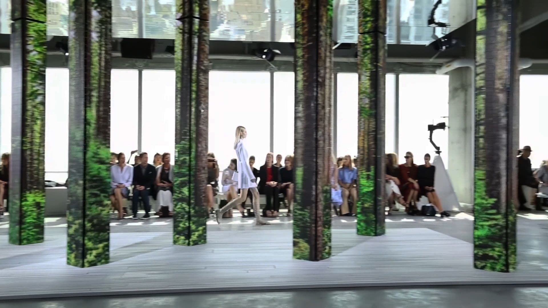 Model in a white outfit walking the runway past seated audience members between forest-print LED columns.