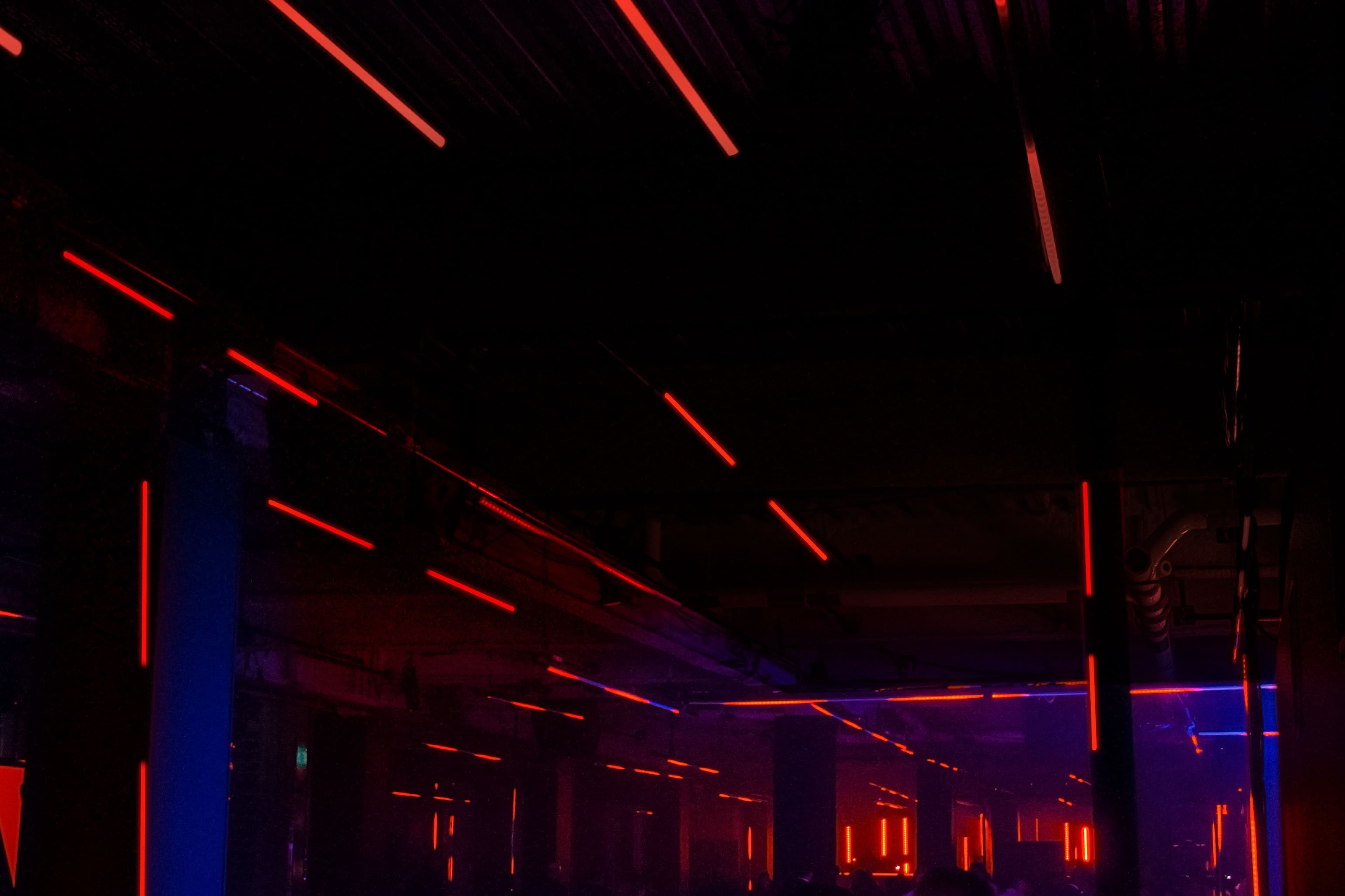 Industrial ceiling and columns crossed by diagonal red neon light beams in a dark venue.