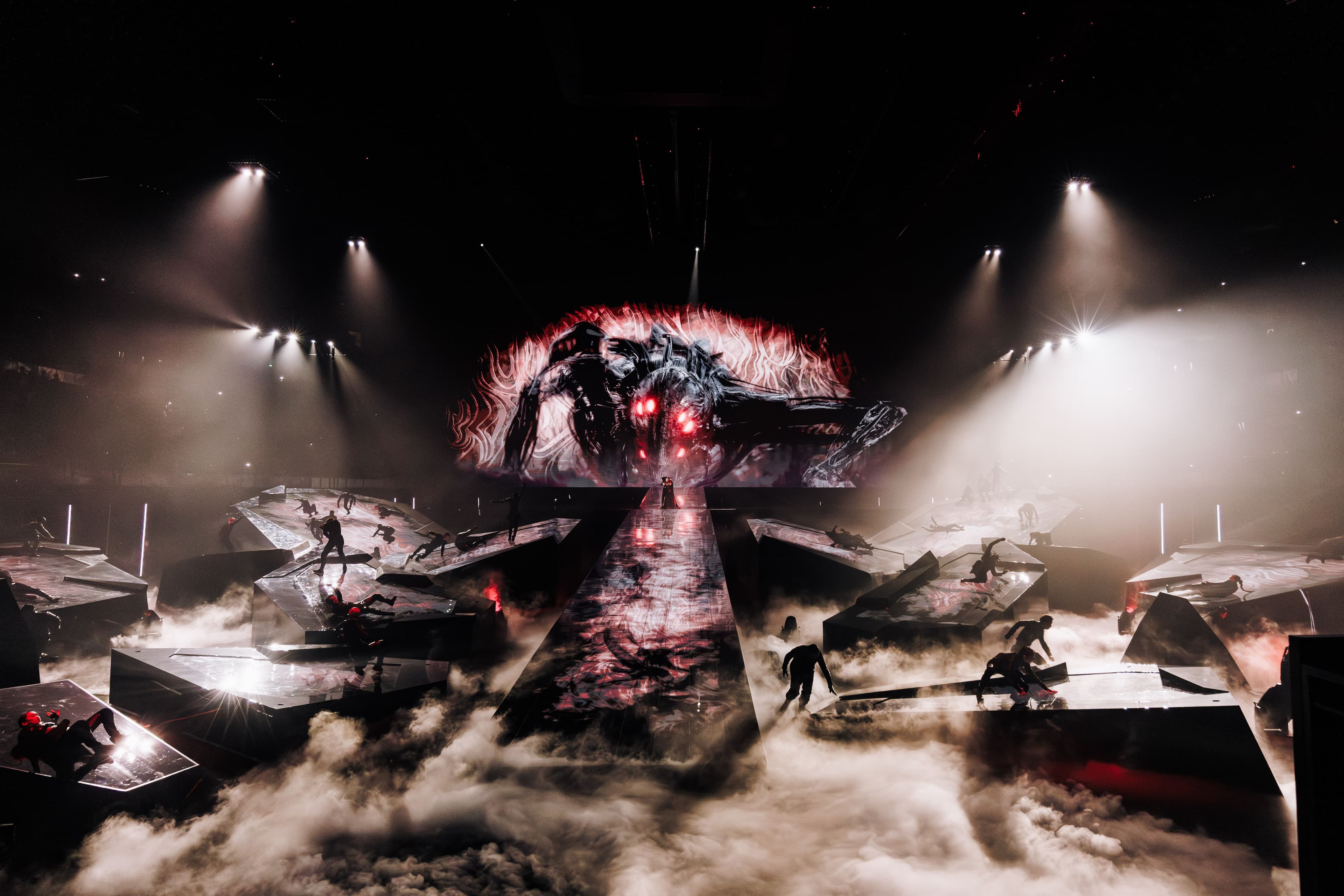 Wide shot of a concert stage with a pyramid-shaped set structure, smoke, and stage lighting, with performers visible on stage.