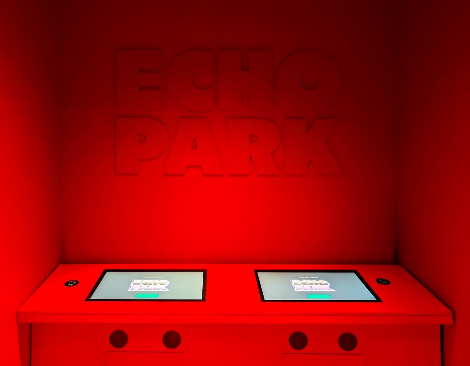 A dark red room with the words Echo Park glowing on the wall above two illuminated touchscreen kiosks on a red console.