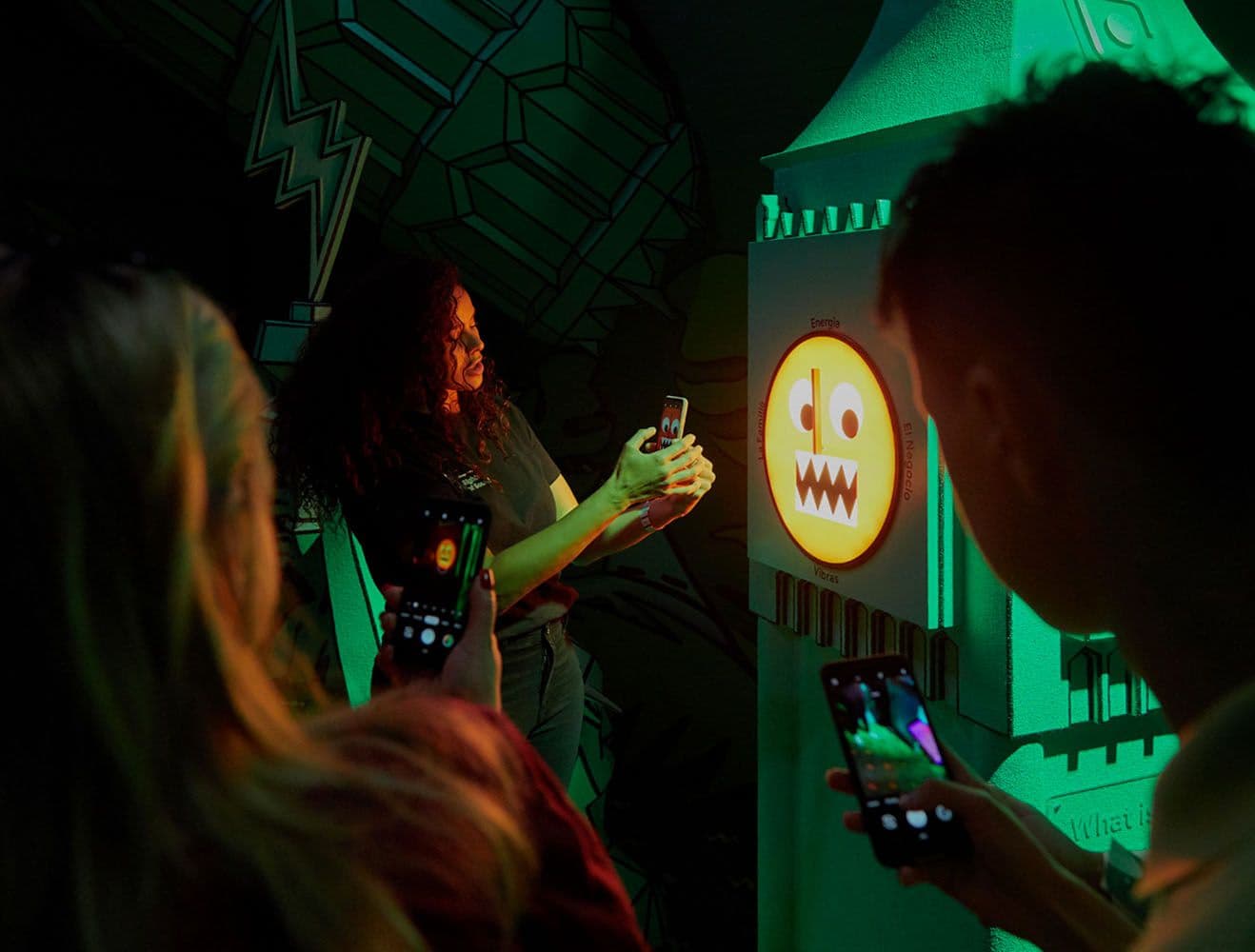 Two people photographing a glowing orange emoji face in a green-lit tropical installation.